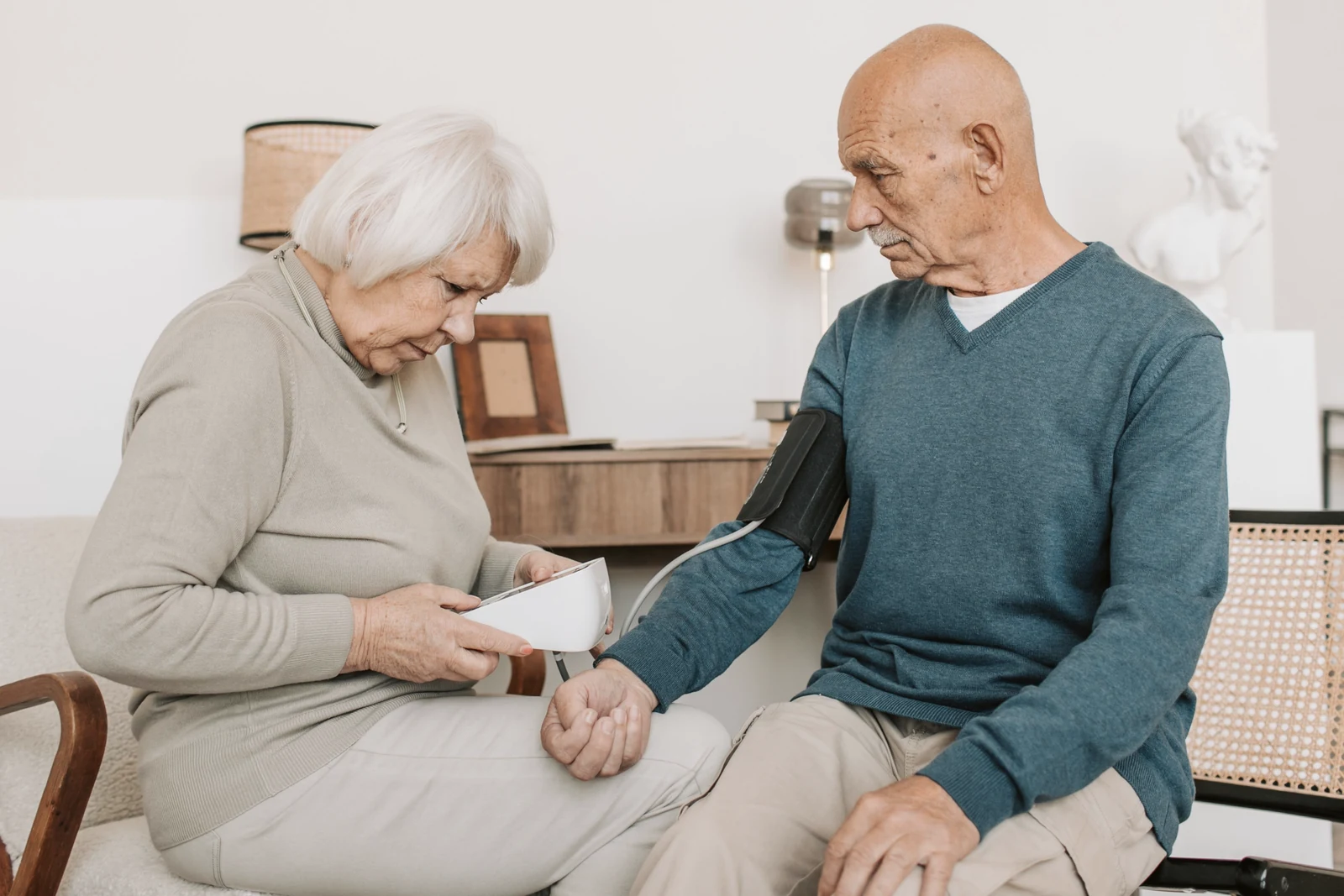 An elderly couple sitting together at home, smiling warmly while checking blood pressure, in a calm reassuring health-monitoring moment