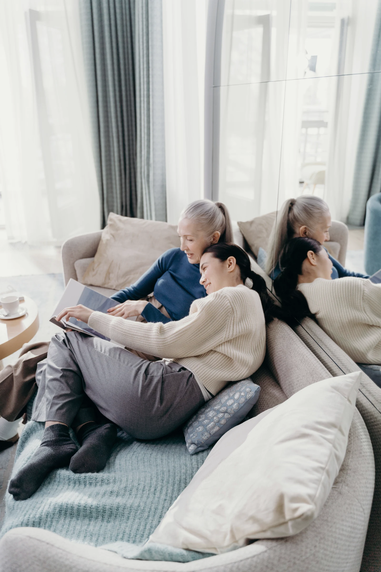 A senior mother and her adult daughter smiling together while reading through a magazine on a sofa at home, a calm and unhurried moment